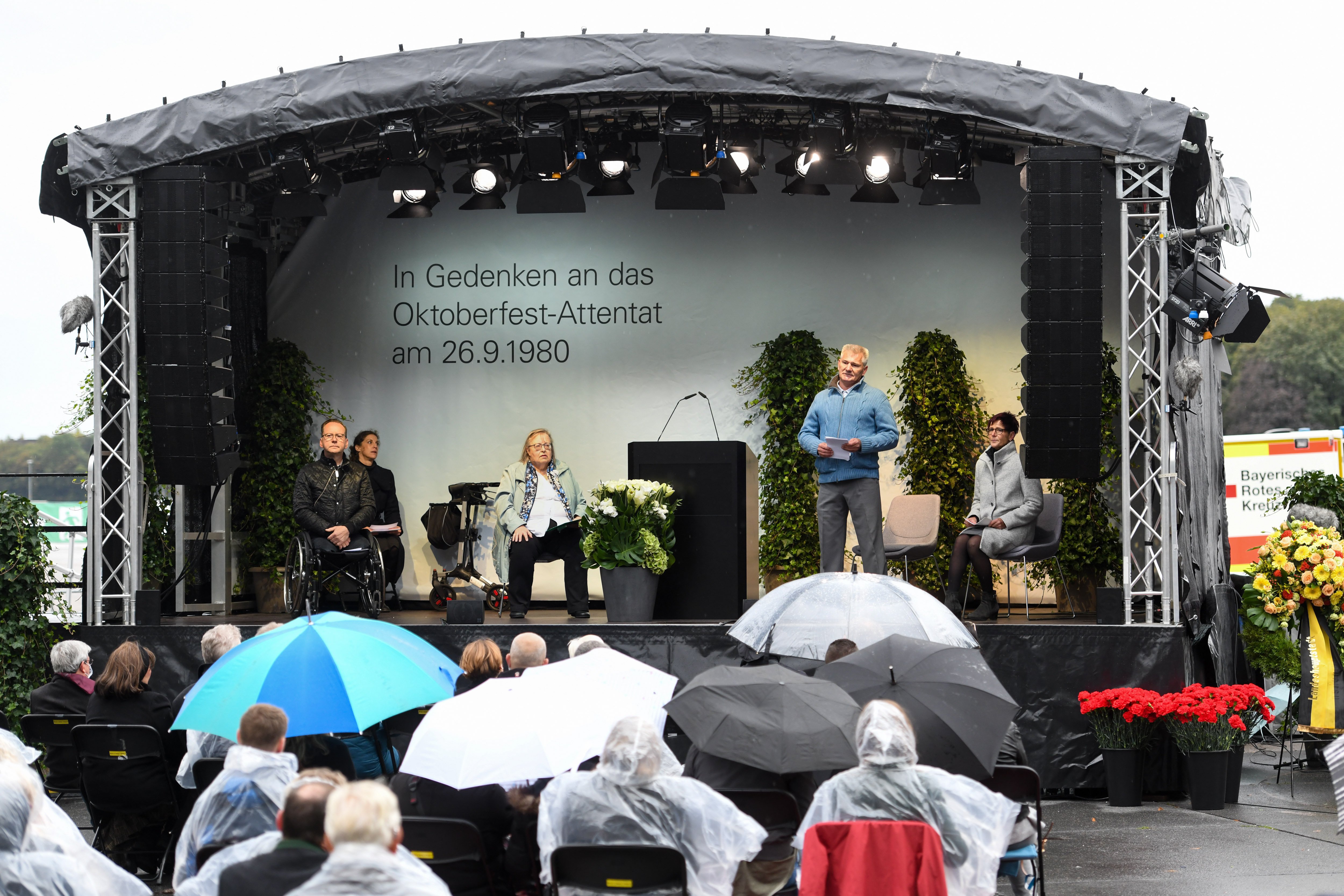 


                 Eine große Bühne auf der Theresienwiese mit merheren Rednern. Vor der Bühne sitzen Zuschauer mit Abstand und Regenschirmen.