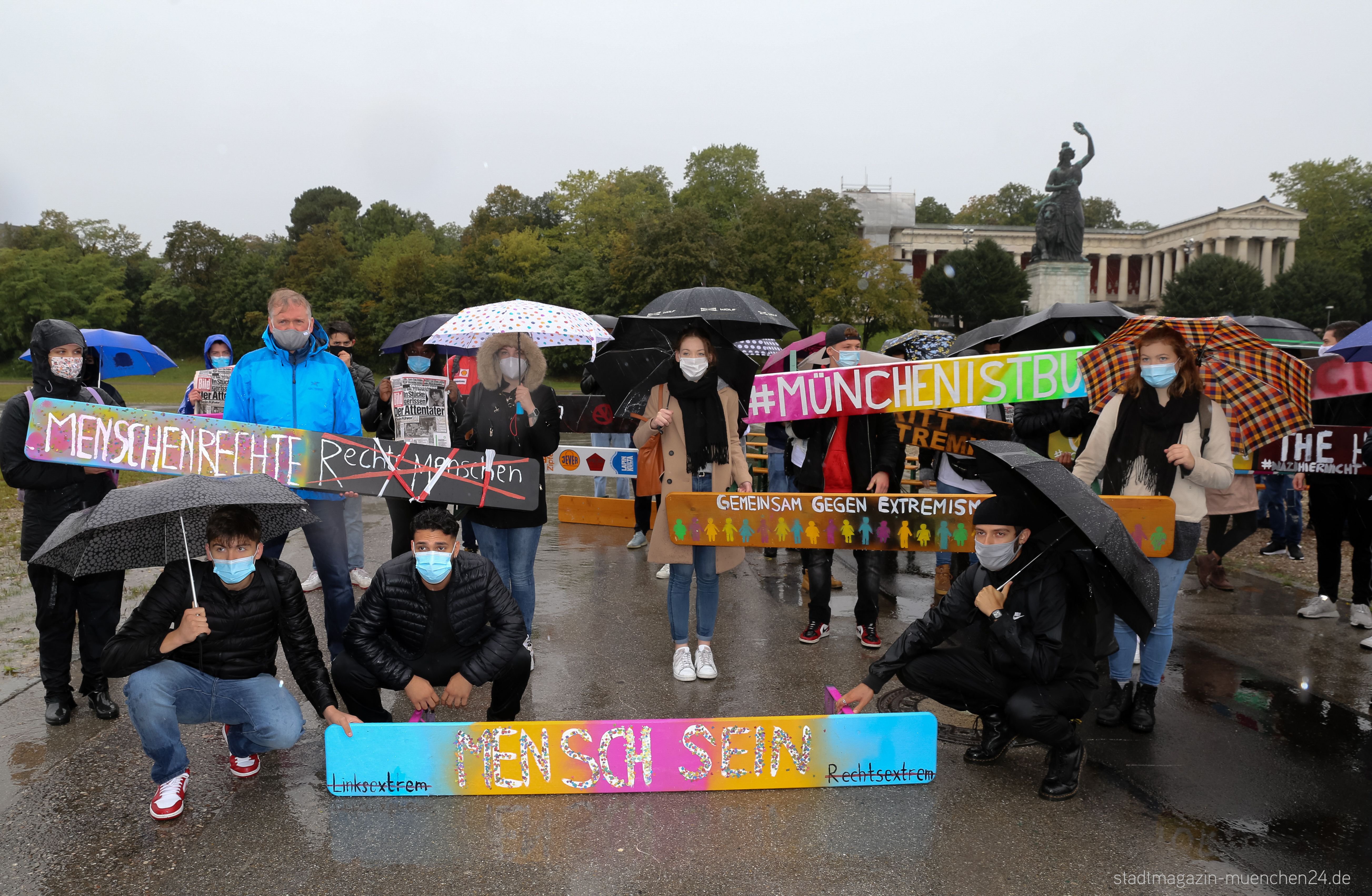 


                 Schülerinnen und Schüler stehen mit Mund-Nase-Bedeckungen und Regenschirmen auf der Theresienwiese und präsentieren bemalte Bierbänke.
