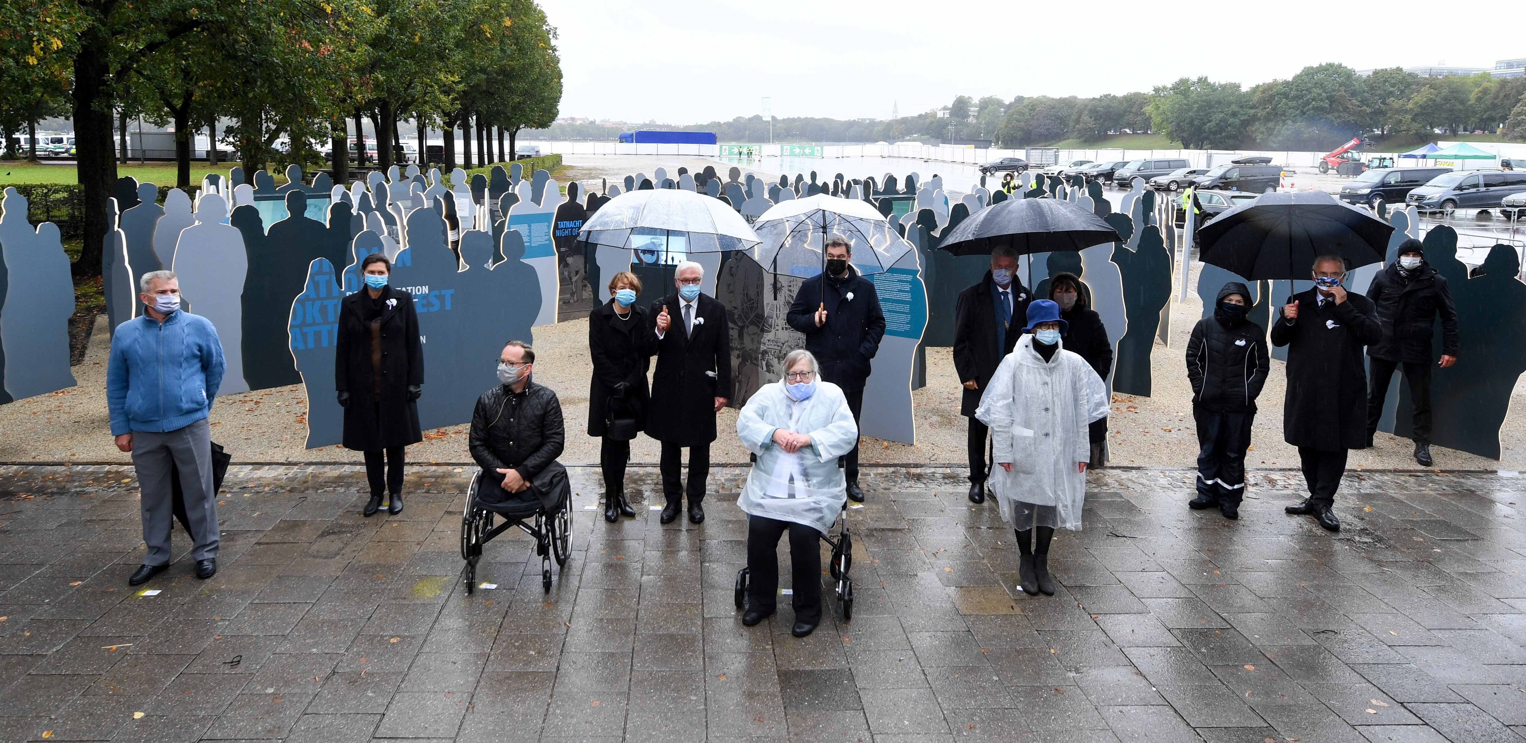 


                 Vier Überlebende und acht Politiker stehen mit Abstand und Regenschirmen vor der Dokumentation des Oktoberfest-Attentats auf der Theresienwiese.