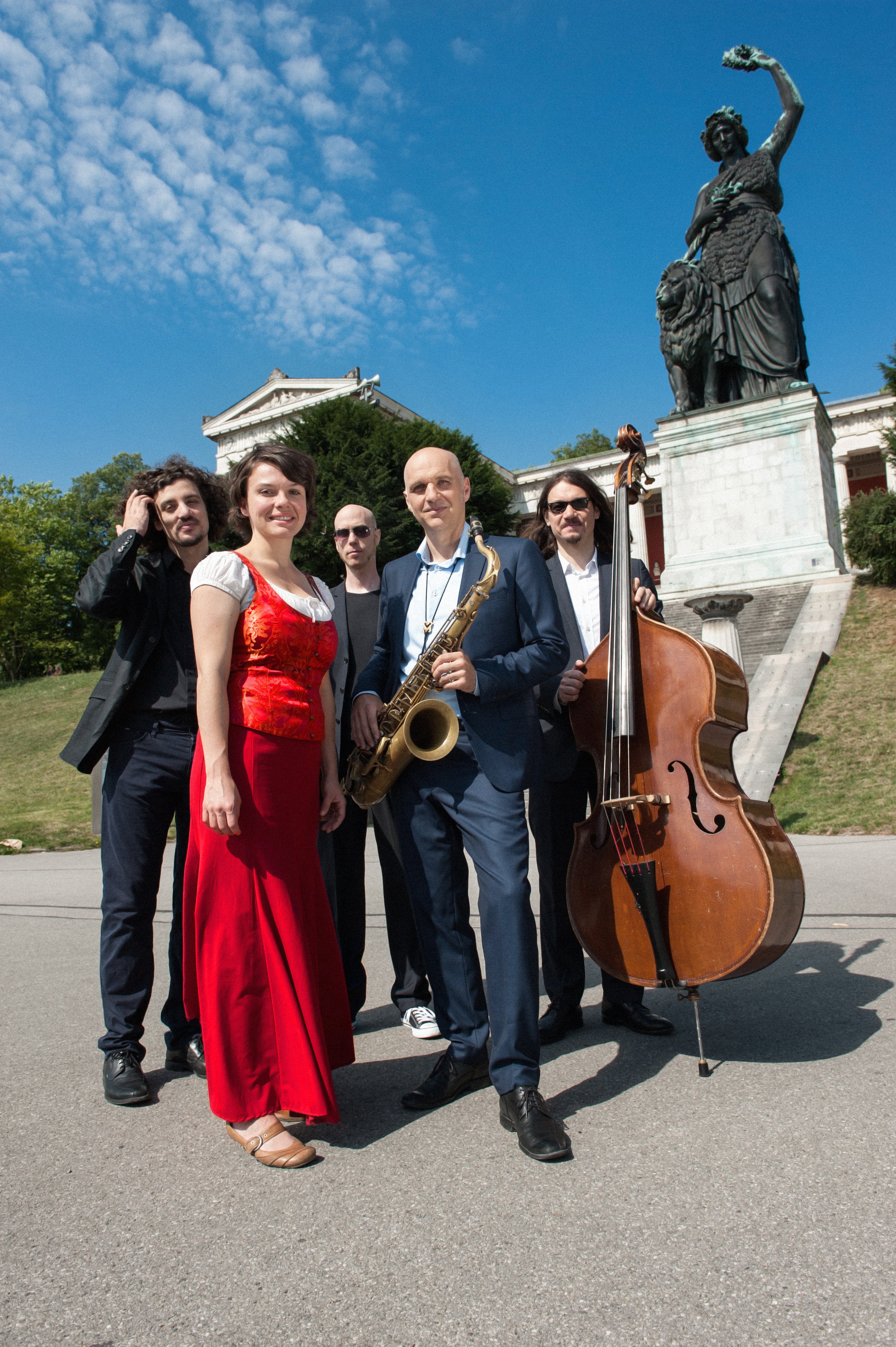 


                 Vier Männer im Anzug und eine Frau im roten Kleid stehen auf der Theresienwiese vor der Bavaria Statue. Der Mann in der Mitte hält ein Saxophon, der Mann rechts außen einen Kontrabass.