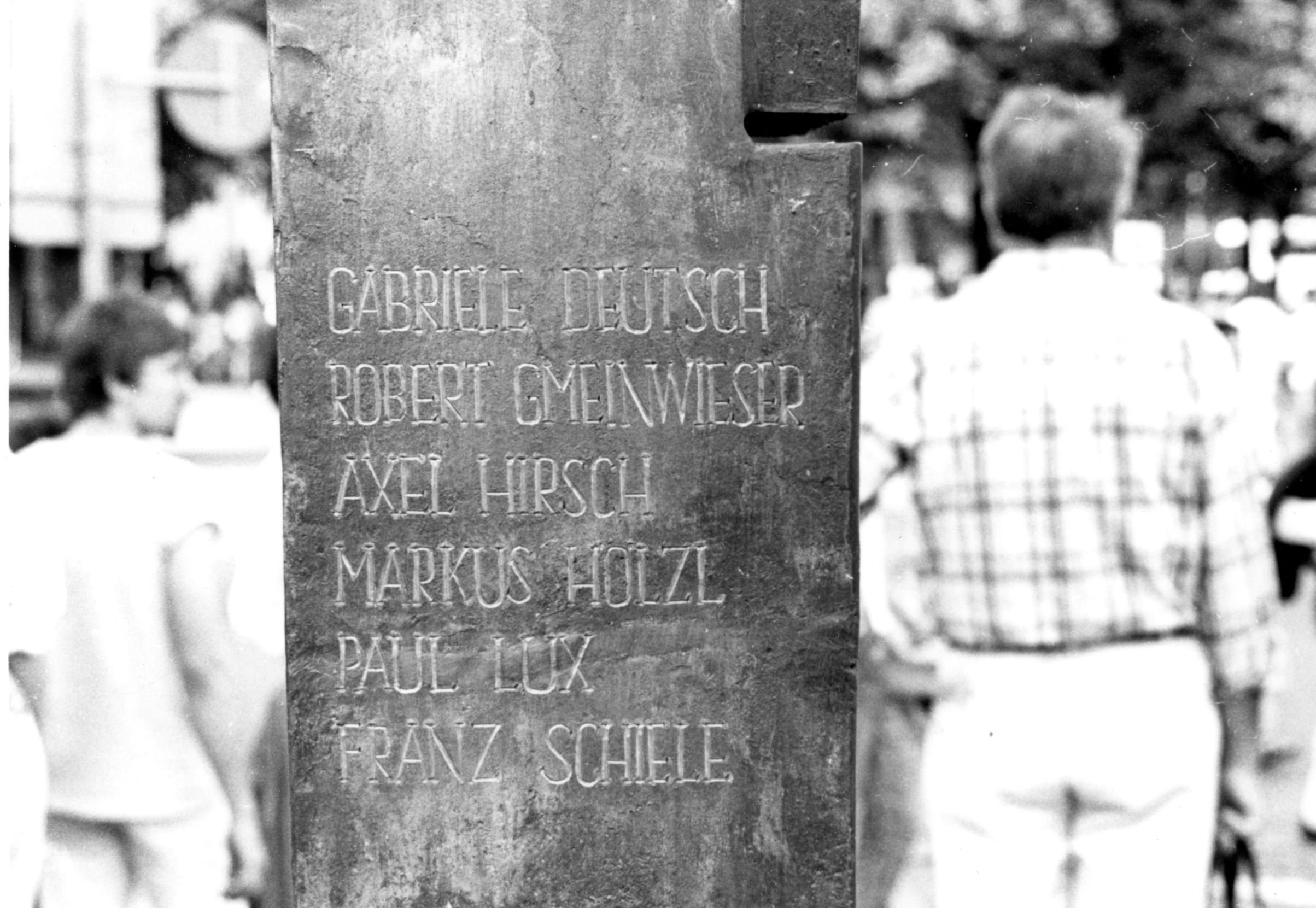 


                 Großaufnahme der Säule mit sechs eingravierten Namen der Todesopfer. Im Hintergrund sind mehrere Personen zu sehen.