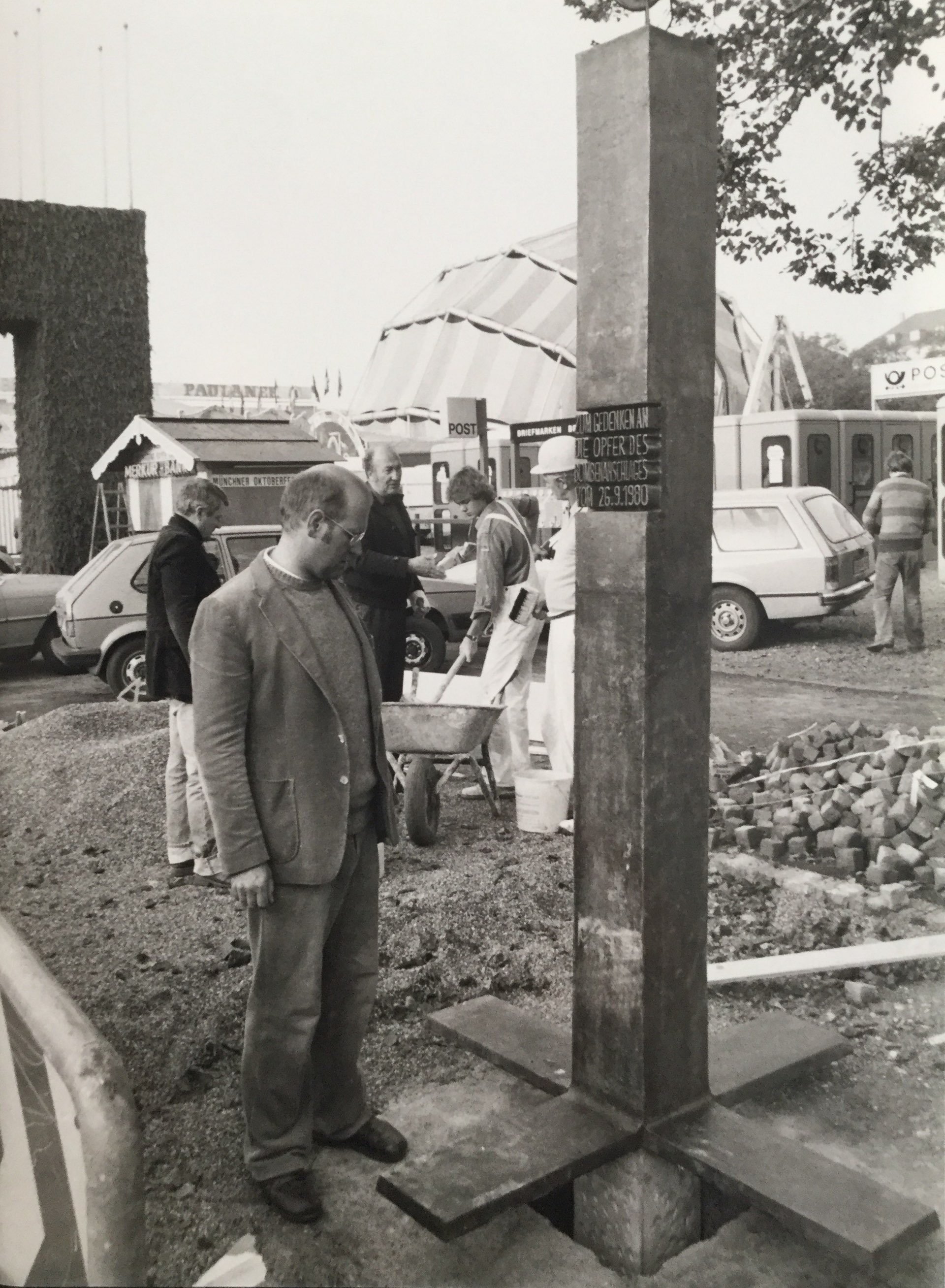 


                 Der Bildhauer Friedrich Koller steht bei der Errichtung des Denkmals 1980 neben der Bronzestele. Weiter im Hintergrund sind Bauarbeiten am Denkmal und für das Oktoberfest zu beobachten.
